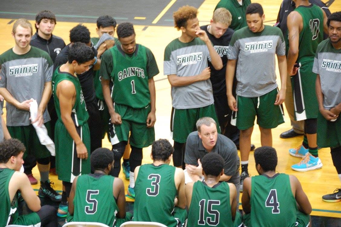 A basketball coach kneels courtside to talk to his players on the bench.