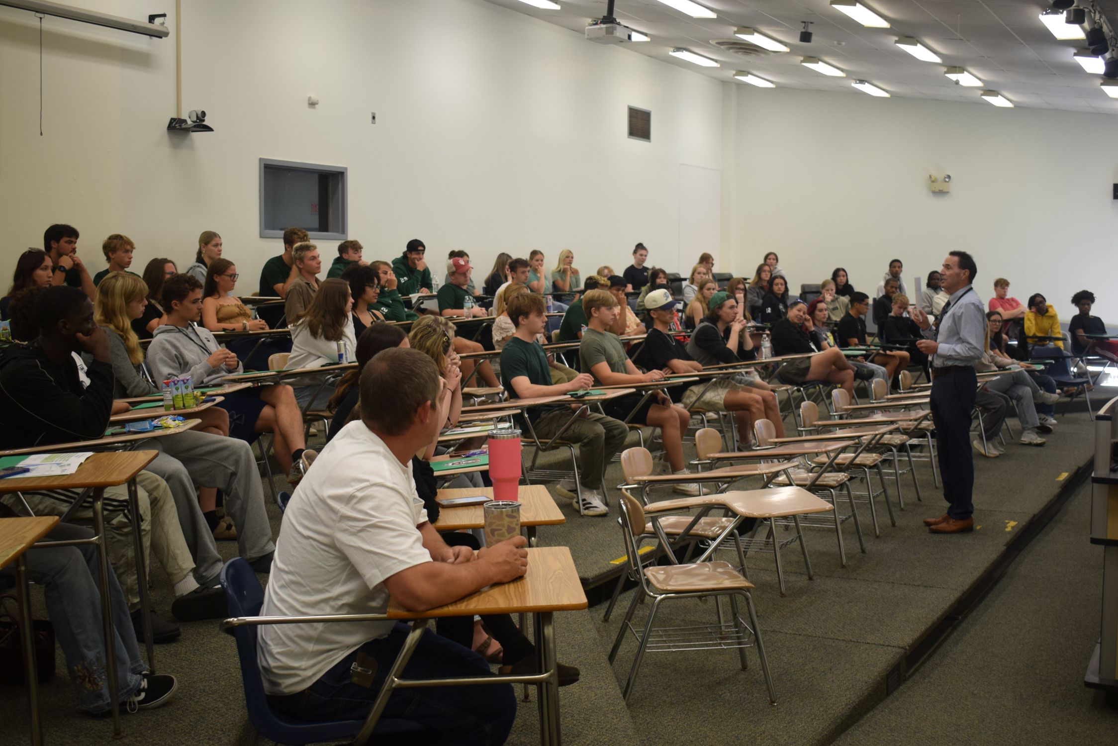 A college president stands in a lecture hall in front of a large group of students