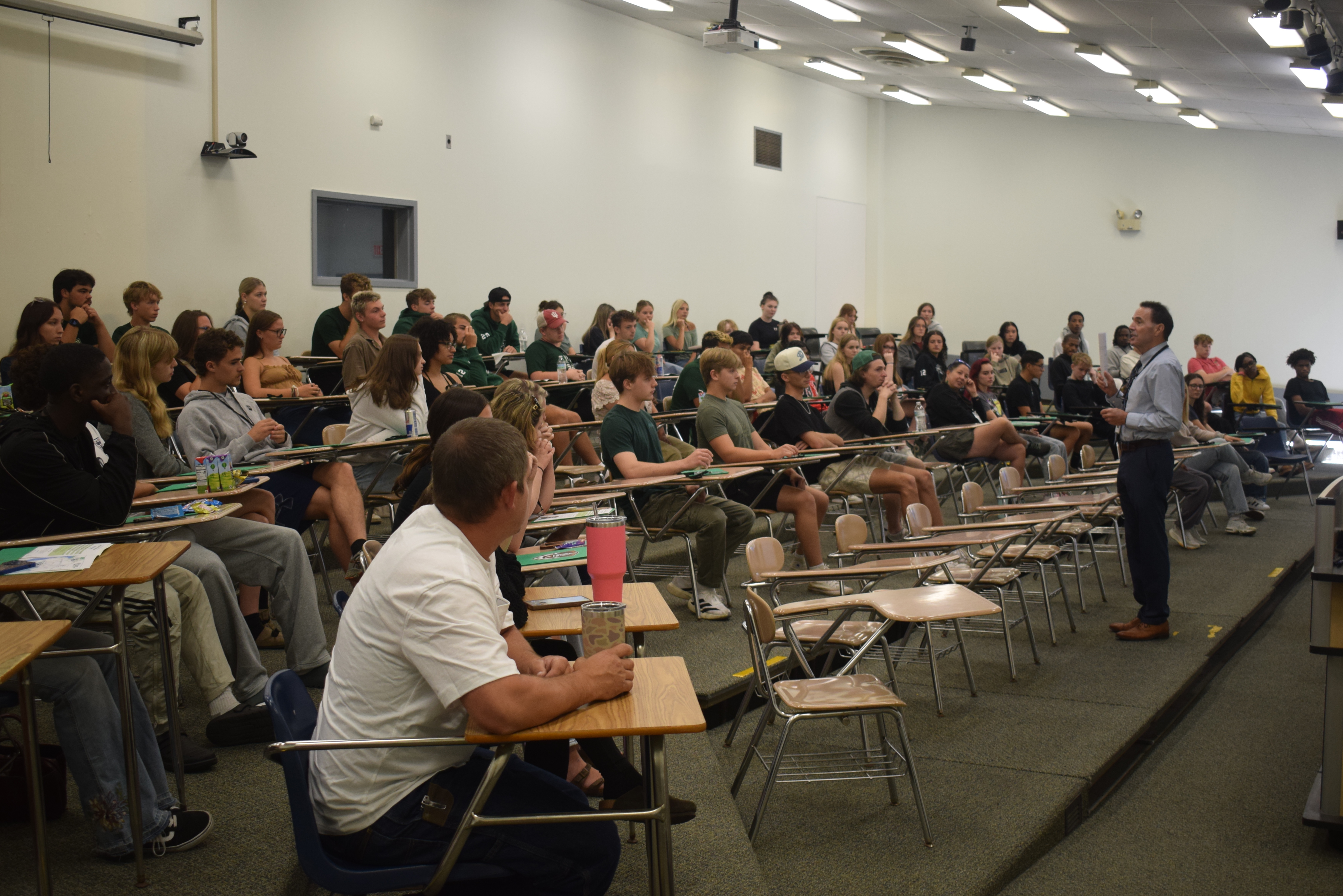 A college president stands in a lecture hall in front of a large group of students