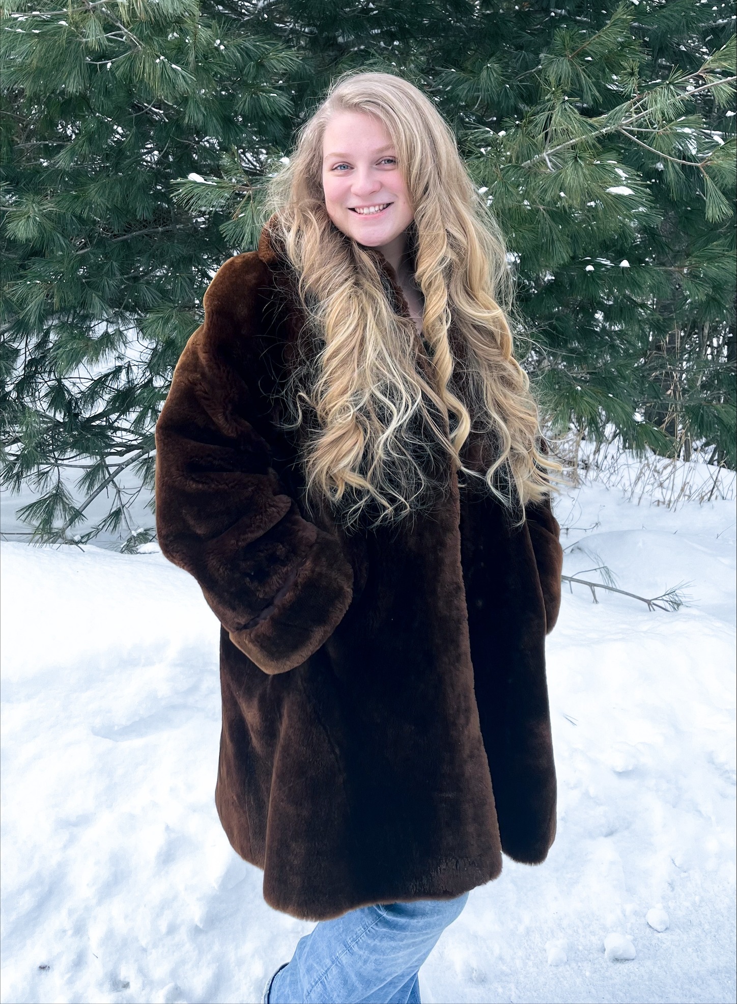 A woman in a fur coat stands in front of a background of trees and smiles at the camera