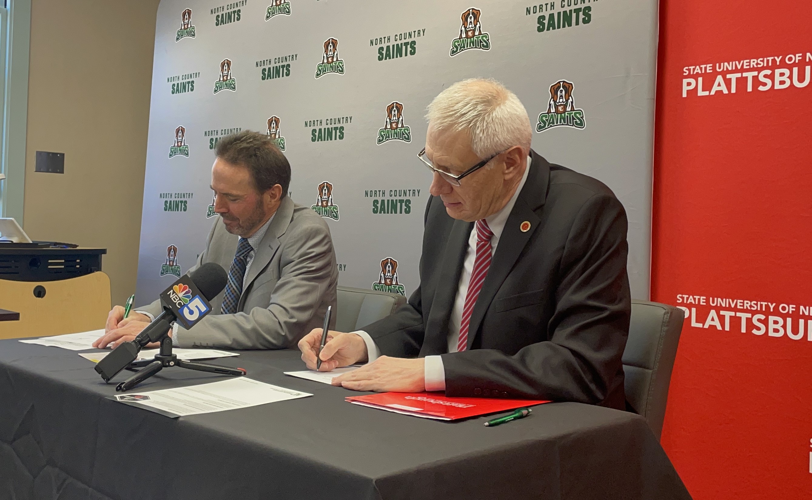 Two college officials sit at a table signing documents in a classroom