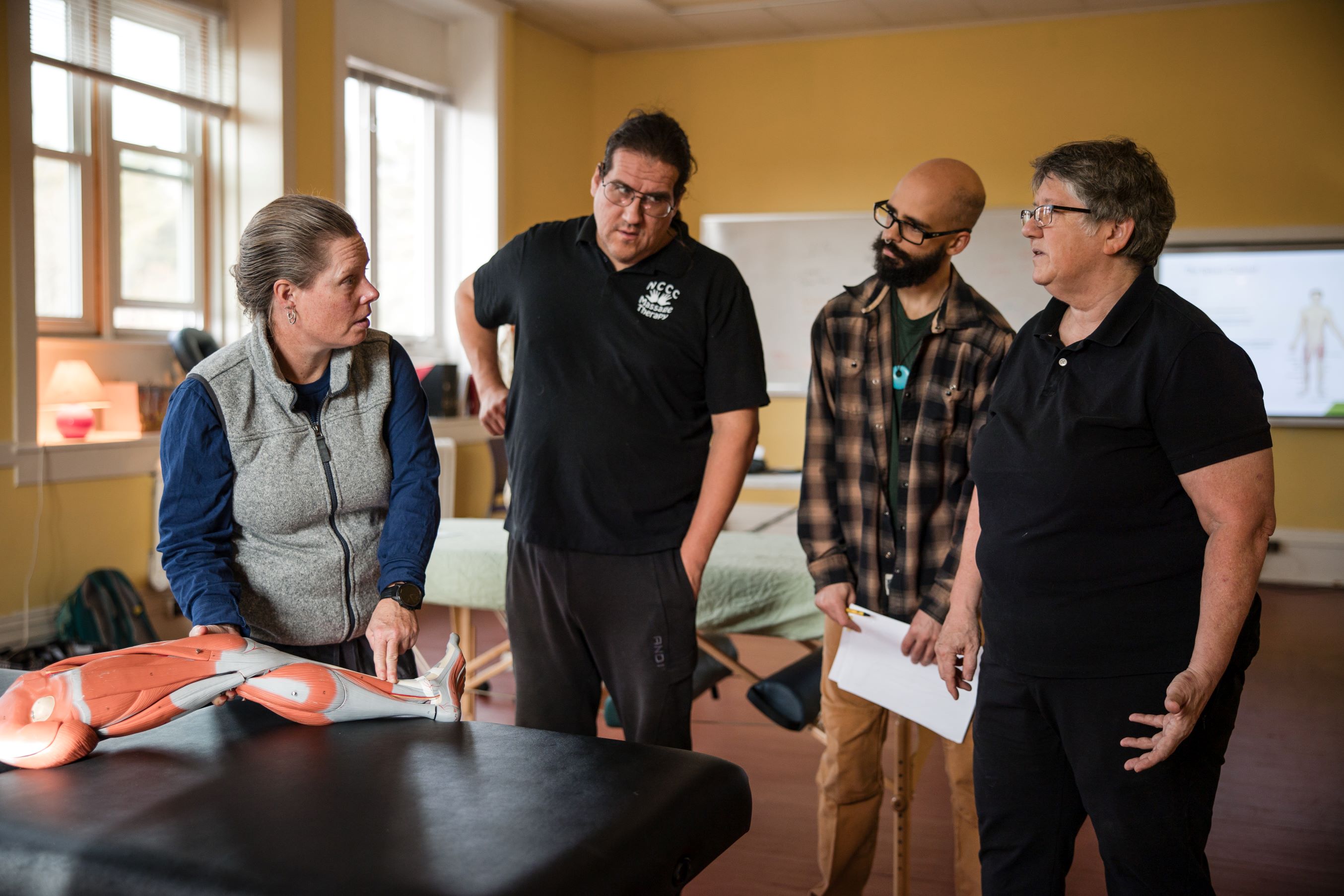 Students stand next to a massage table and talk with an instructor in a classroom.