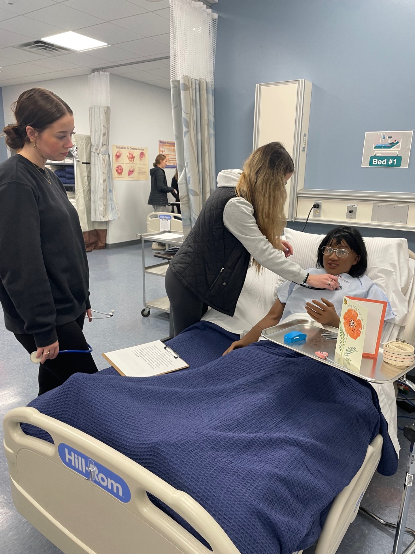 Two high school students stand next to hospital bed looking at a nursing mannequin