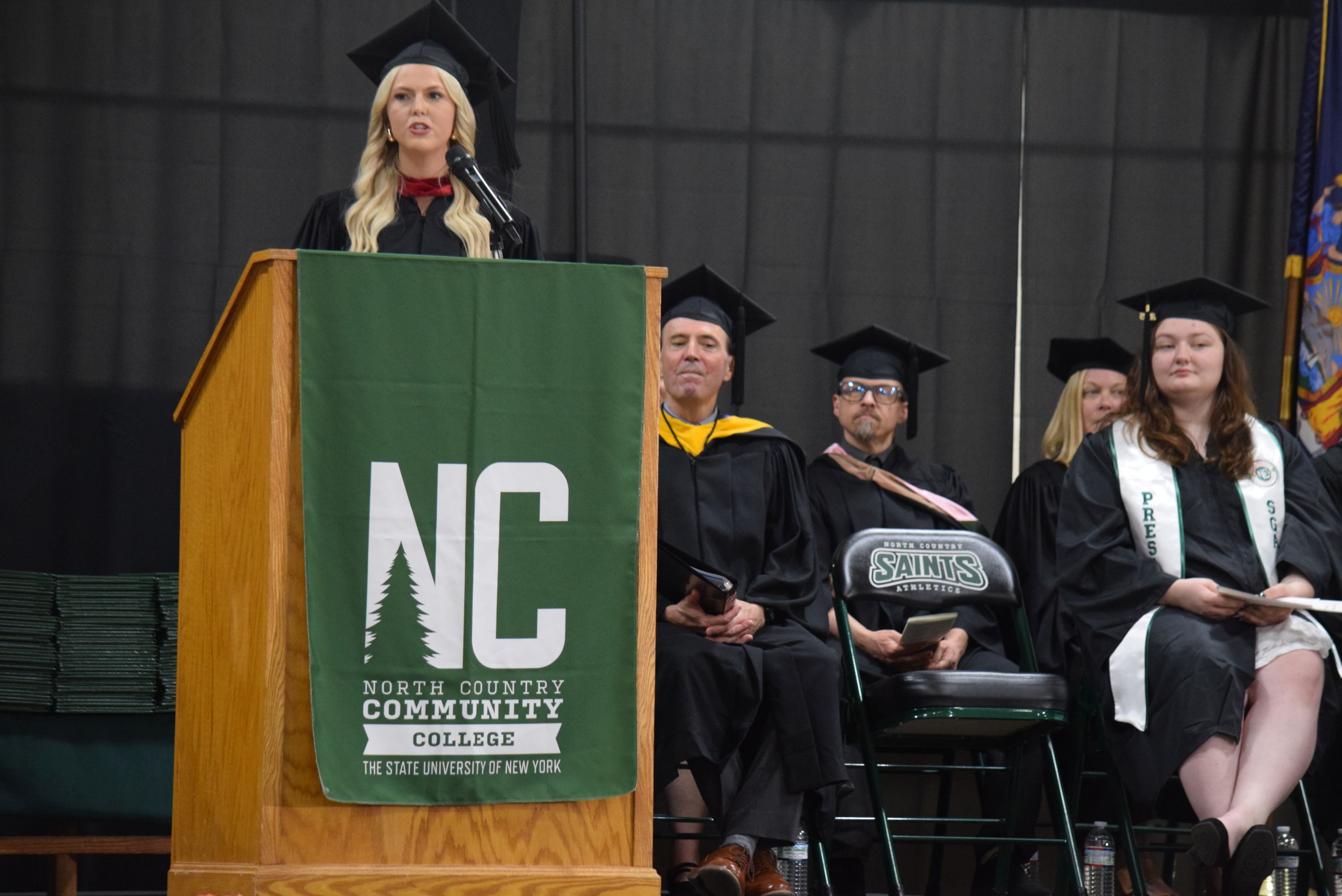 A speaker stands at a podium on a stage speaking to an audience of college graduates.